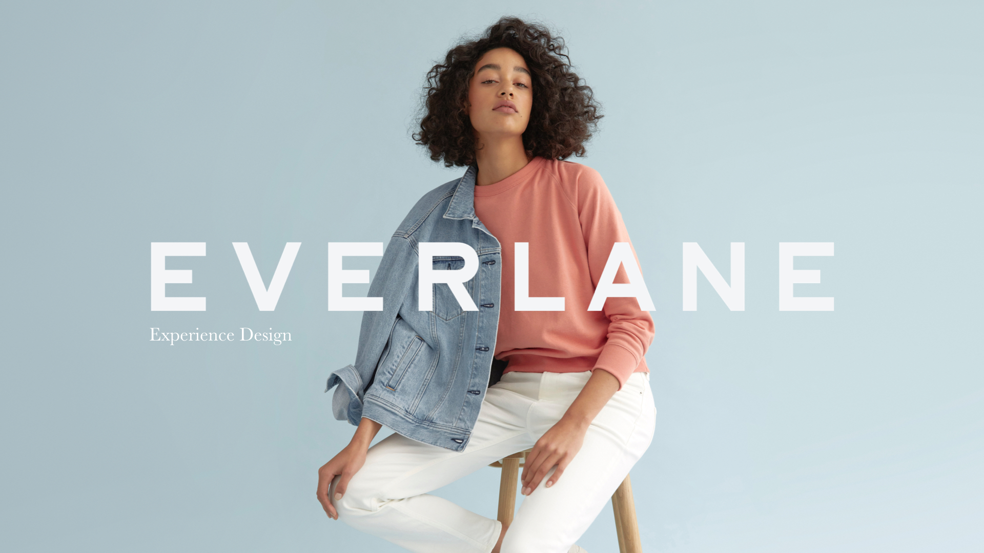 Logo d'Everlane au centre en avant-plan. À l'arrière, une femme habillé d'une veste en jean, d'un pull rose et de pantalon blanc.